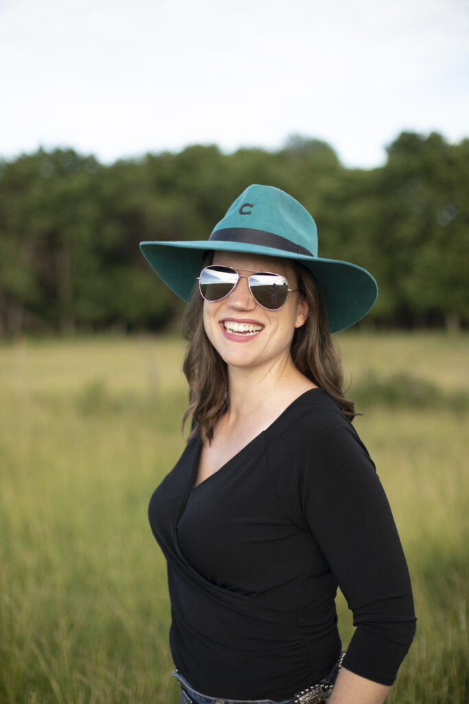 Woman with brown hair in a teal cowboy hat and aviator sunglasses